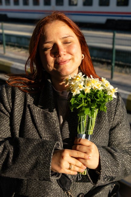 A young woman with red hair, eyes closed and a gentle smile, is holding a bouquet of fresh yellow and white daffodils with green foliage. The bouquet is wrapped in clear glass, and she is wearing a da