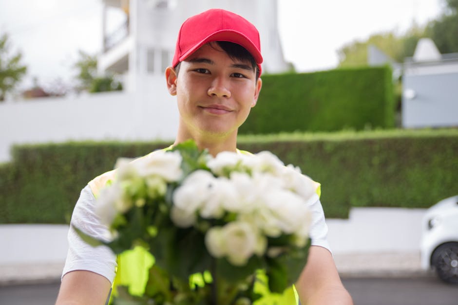 A delivery person wearing a red cap and high-visibility vest is standing outdoors on a residential street. They are holding a fresh floral bouquet with white flowers and green foliage, extending it to