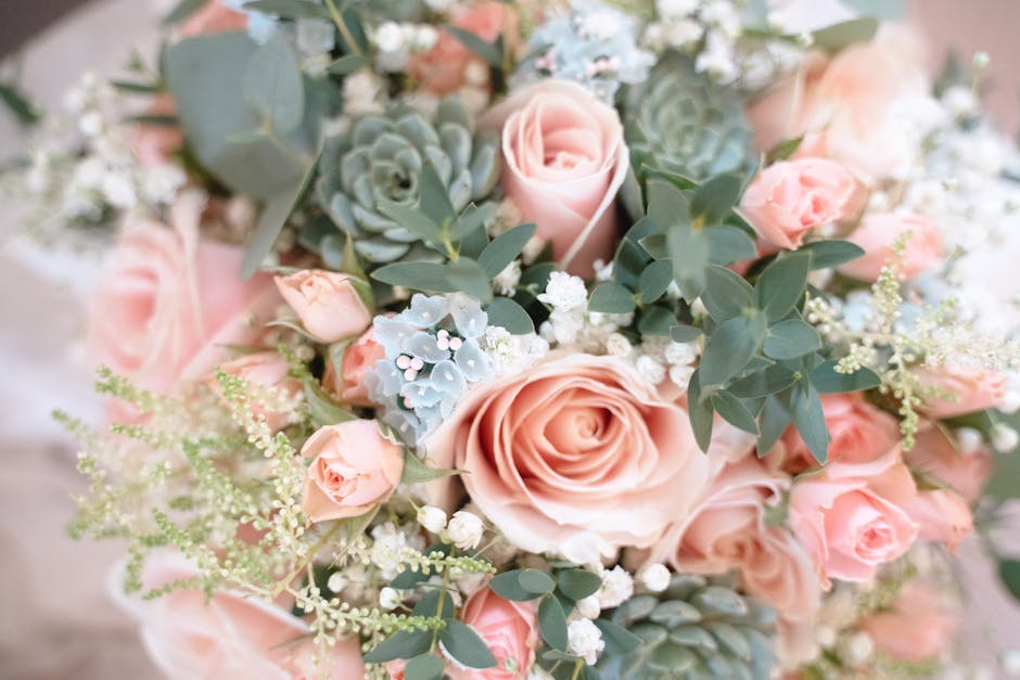 A close-up image of a floral bouquet featuring large, soft pink roses in full bloom, complemented by smaller pink rosebuds, delicate white baby's breath, silvery-green eucalyptus leaves, and blue-gree