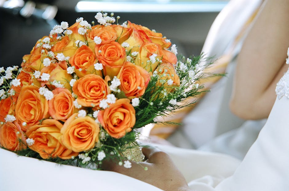 A close-up of a bouquet arranged with vibrant orange roses intertwined with small white filler flowers, accented by delicate greenery. The bouquet is wrapped in elegant transparent cellophane with a s