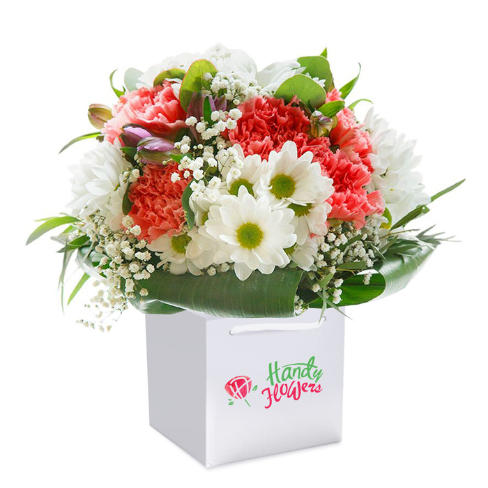 Presented upright within a crisp white square gift bag, this expertly arranged bouquet captures the essence of Balham's vibrant community spirit. The arrangement features plush coral and deep red carnations, their textured, frilled petals layered with precision for a sense of abundant fullness. Bright white daisy-like flowers with chartreuse centers bring uplifting contrast, their clean lines reminiscent of the fresh start to a weekend morning in Balham. Cascading throughout, delicately branching baby's breath lends an ethereal softness, entwined among the primary blooms to create depth and visual intrigue. Discreet touches of purple-hued buds nestle on the bouquet's left, offering subtle variation and anticipation of petals soon to unfurl. The bouquet is surrounded by lush, broad-leaved greenery that forms a natural collar, echoing the mature parks and gardens dotted across the neighbourhood. The white gift bag-designed for both elegance and practicality, with robust ribbon handles-anchors the arrangement and hints at the convenience of same-day delivery from Balham Flowers. The polished white background enhances every detail-the lush textures, vivid tones, and meticulous floral artistry. This visually captivating floral gift is ideal for celebrating special occasions in SW12, whether it's an anniversary, birthday, or simply expressing affection to a loved one, and stands as a testament to Balham's proud tradition of quality local floristry.