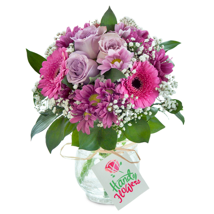 A richly textured floral bouquet, overflowing with uplifting colour and crafted with Balham's creative energy, makes a striking impression set against a crisp white background. At the heart of this lush display, several elegant lavender roses unfurl with stately grace, their soft, cool tones setting a calm yet joyful mood. Surrounding these are jubilant hot pink Gerbera daisies, their broad petals and contrasting dark centers instantly catching the eye-reminiscent of the energetic vibe along Hildreth Street Market. Accentuating the arrangement's depth, velvety magenta chrysanthemums are clustered throughout, each bloom with intricate, layered petals and hints of yellow-green at the center, offering subtle surprises and visual interest. Interspersed with clouds of delicate white baby's breath, the bouquet feels light and full of movement, as if swaying in a gentle Balham breeze. Glossy dark green foliage peeks from beneath and frames the blossoms, grounding the colours with natural harmony. Housed in a clear glass vase with a distinctive cracked texture, the flowers are secured by a neat raffia tie, from which a birthday tag with bold red and green script and a miniature flower illustration dangles with a festive wink. This handcrafted bouquet, perfect for delivery throughout Balham and nearby SW12, is ideal for birthdays, heartfelt thanks, or brightening home and heart with authentic local charm.